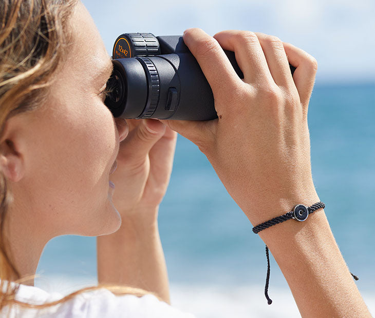 The 4ocean Osborne Reef Bracelet is worn by a female model at the beach as she uses binoculars.