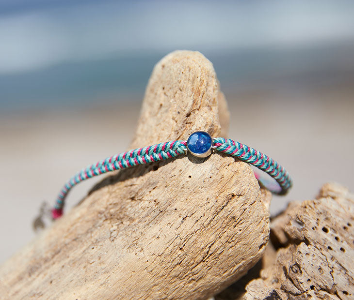 4ocean Ocean Drop Bracelet. Pink, green and teal braided cord with stainless and blue bezel being displayed on a rock with the beach in the background