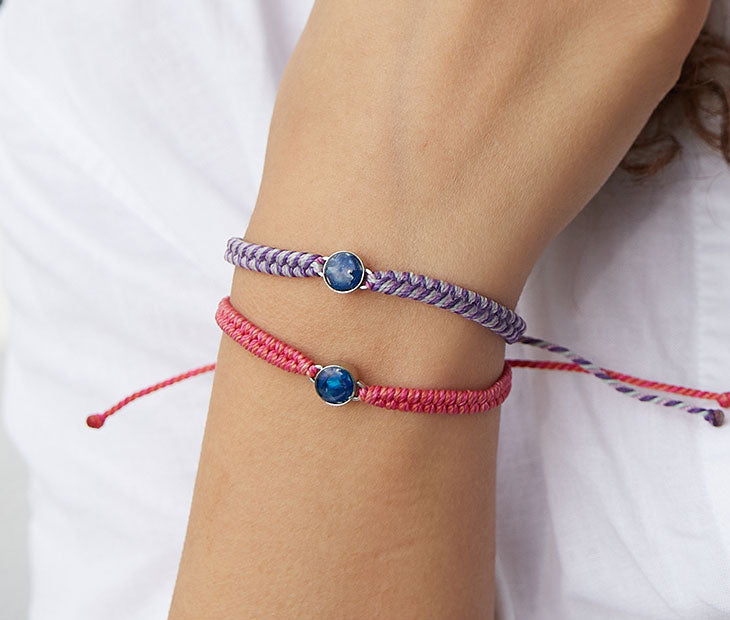 4ocean Pink Ocean Drop Bracelet. Pink braided cord with stainless and blue bezel. Shown on female model with a coordinating 4ocean Purple Ocean Drop Bracelet.
