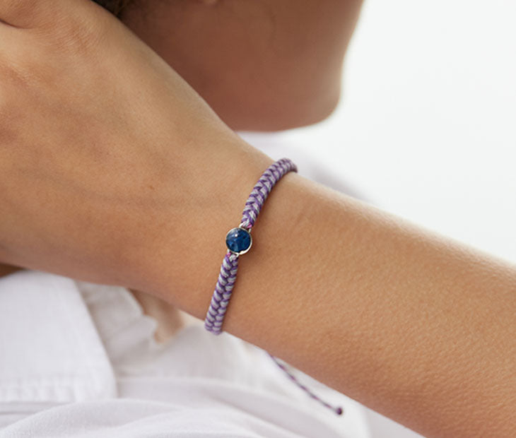 4ocean Purple Ocean Drop Bracelet. Purple braided cord with stainless and blue bezel. Shown on female model.